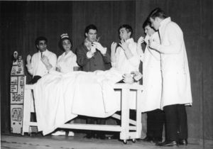 Suffolk University students perform onstage for a theater production, dressed in white medical lab coats