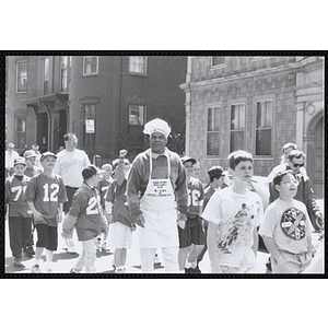 A male staff member wearing a chef hat and an apron embroidered with the text "Charlestown Boys and Girls Club 25 Yrs. and Still Cooking," walking in the Boys and Girls Clubs of Boston 100th Anniversary Celebration Parade