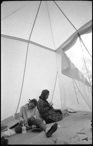 Inside of a strikers' tent, two young women seated on the ground, drinking milk through a straw