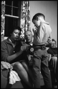 Mildred Loving seated on a couch, fixing a toy cowboy belt and holster onto her son; Richard Loving looks on