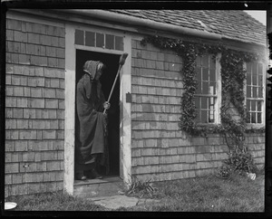 Reuben Austin Snow, the cross-dressing hermit of Cape Cod, wearing a shawl, wielding a broom, and standing in the cottage doorway