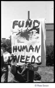 Protester at a Mobilization for Survival antinuclear demonstration near Draper Laboratory, MIT, with sign reading 'Fund human needs: No more Hiroshimas'