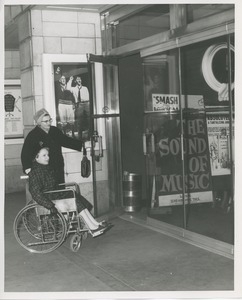 Mrs. Frances Marsala seated in her wheelchair entering a theater with a friend or attendant