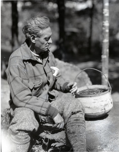 George Allan England, seated on a rock with cooking pot