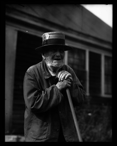 Older man, age 101, wearing a well-weathered hat, leaning on the handle of a hoe