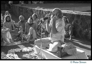 Ram Dass seated on a small platform, arms raised, during his appearance at Andrews Amphitheater, University of Hawaii