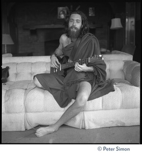 Krishna Das, playing bass guitar, sitting on a couch in the Vicente Street house