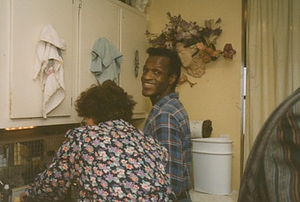 A Photograph of Marsha P. Johnson in a Yellow Kitchen Wearing a Blue Flannel Shirt, Smiling Over Her Shoulder