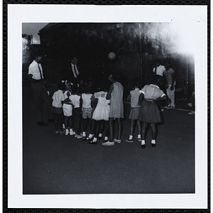 Eight girls, facing away from the camera, stand in a line in front of two male staffers and look at something during a Roxbury Boys' Club event