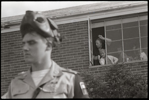 Antiwar demonstration at Fort Dix, N.J.: military policeman with gas mask raised, woman and children in window behind flashing peace sign