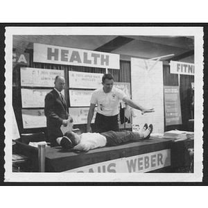 Young woman lies on a table during a fitness test, in front of a banner reading "health" while two men stand nearby, one of them gesturing to her