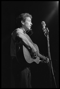 Bob Dylan, with guitar and harmonica, performing on stage, Newport Folk Festival