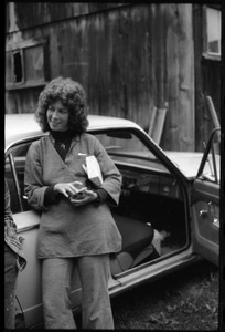 Unidentified woman standing by a car with open door in front of the barn, Montague Farm commune