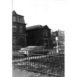 View of La Alianza Hispana's headquarters, 409 Dudley Street (building on left) and 407 Dudley Street (on right), taken from across the street; there is a fence and cars parked in the foreground.