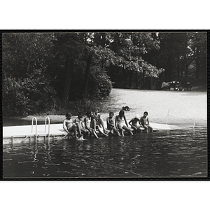 Seven youth and a supervisor sit on a dock with their legs in the water