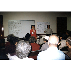 Woman teaches at the front of a classroom, while Association members sit listening