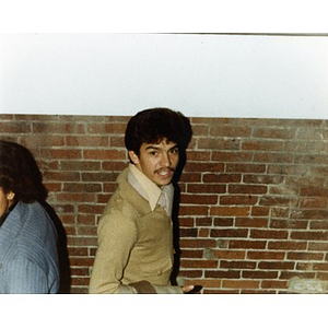 Hispanic American male employee wearing a beige sweater standing in front of a brick wall at a Christmas party at La Alianza Hispana offices, Roxbury, Mass