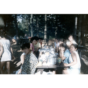 Young adults and children sit in the woods at a picnic table covered with food
