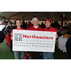 Ginny Driscoll, Marion Stanley, Vice President for University Corporate Partnerships, and unknown hold up a placard for the NU Corporate Partnerships table during the Homecoming game