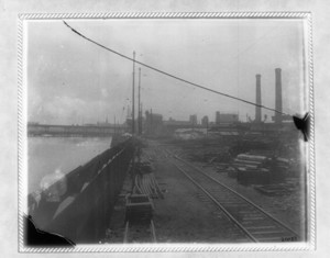 View of tracks by the waterfront with smoke stacks in the background to the right