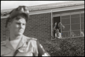 Antiwar demonstration at Fort Dix, N.J.: military policeman with gas mask raised, woman and children in window behind flashing peace sign