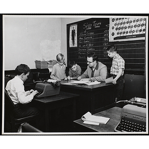 A man tutors three boys in a typewriting class as another boy uses a typewriter at a desk