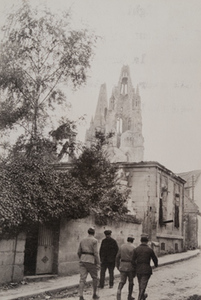 3 soldiers and one civilian walk along a street in the town of Soissons, a damaged church steeple in the background