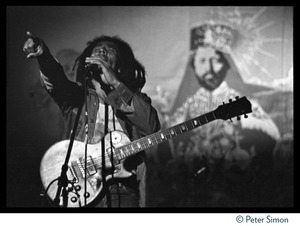 Bob Marley in concert, exhorting the audience while standing in front of a large banner with a portrait of Haile Selaisse