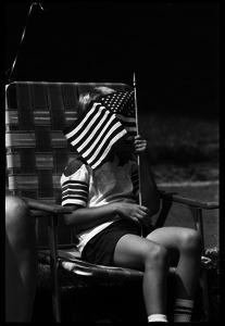 Boy in a lawn chair, ducking behind a small American flag while watching the Chesterfield's Fourth of July parade