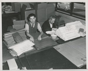 Two men, one with cruches, fold and roll package wrappers at a sheltered workshop