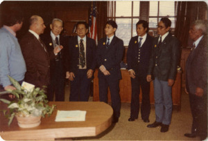 Photograph of a group of men standing in an office and listening to a person speak, [1982-1983].