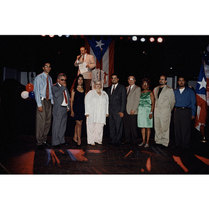 Nine people, including Carmen Pola, stand in front of a stage where a man speaks into a microphone at a Festival Puertorriqueño event