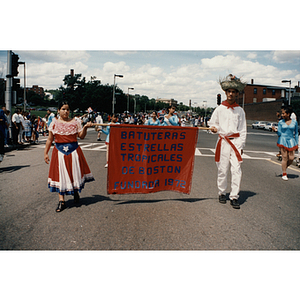 Two people in costume carry a banner for the Estrella Tropicales performing group during the Festival Puertorriqueño parade, near Centre Street and Columbus Avenue