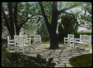 Flagstone patio built around a tree, with white wooden chairs and benches (damaged slide)