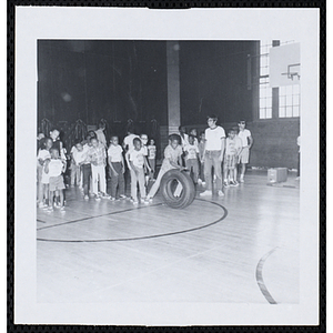 A boy participates in a tire rolling race in a gymnasium as other boys look on during Tom Sawyer Day