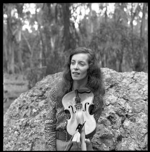 Woman posed with a fiddle, seated on a granite boulder in a eucalyptus grove