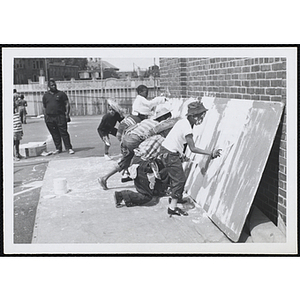 A group of boys apply white paint on large board as a man looks on during Tom Sawyer Day