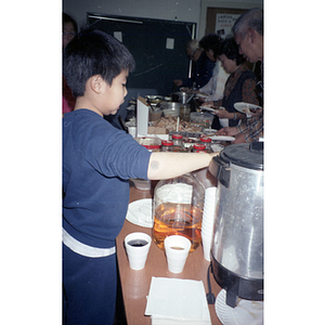 Young boy stands beside a buffet table to serve himself some juice at a Chinese Progressive Association holiday party
