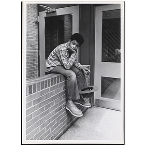 A boy from the Boys' Clubs of Boston sitting in front of a door and resting his head in his hand while holding a strap on his hat