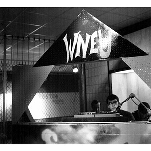 Young man with sound equipment sitting behind a sign reading "WNEU"