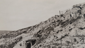 Red Cross worker walking along a war-torn hillside above barbed wire and a dugout entrance
