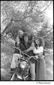 Peter Simon seated on a motorcycle, with Usha (Cathy Brown, right) and two other admiring women caressing him