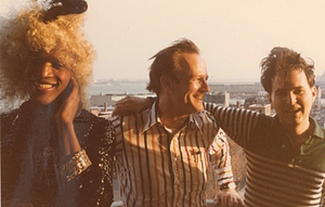 A Photograph of Marsha P. Johnson with Curly Blonde Hair and a Black Sequined Jacket, Posing with Randy Wicker and Another Person