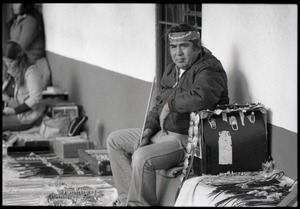Native man seated on a bench, selling bolo ties and concho belts