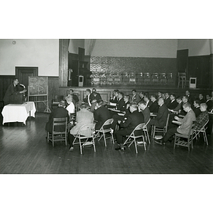 A speaker stands at the podium while staff members, seated in chairs, look on during an administrative meeting