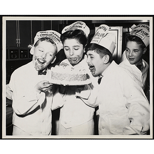 Three members of the Tom Pappas Chefs' Club with frosting smeared on their faces pose with a decorated cake in a Brandeis University kitchen