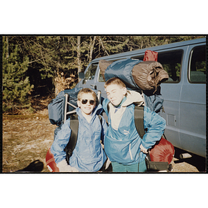 Two boys with large backpacks stand next to a van outdoors