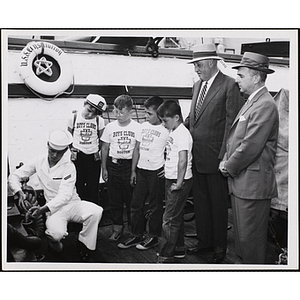 Four boys stand with Boston Rotary Club President Arthur Curren Jr. (far right) and Rotary Club committee member Clement A. Stodder (right) on the deck of the USS Constitution, watching sailor Edward Smith works with an anchor
