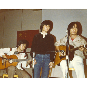 Three Chinese men sing and play guitar onstage at the 30th anniversary celebration of the People's Republic of China held in the Josiah Quincy School auditorium