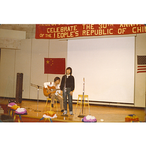 Chinese man sings onstage accompanied by a man playing guitar at the 30th anniversary celebration of the People's Republic of China held in the Josiah Quincy School auditorium